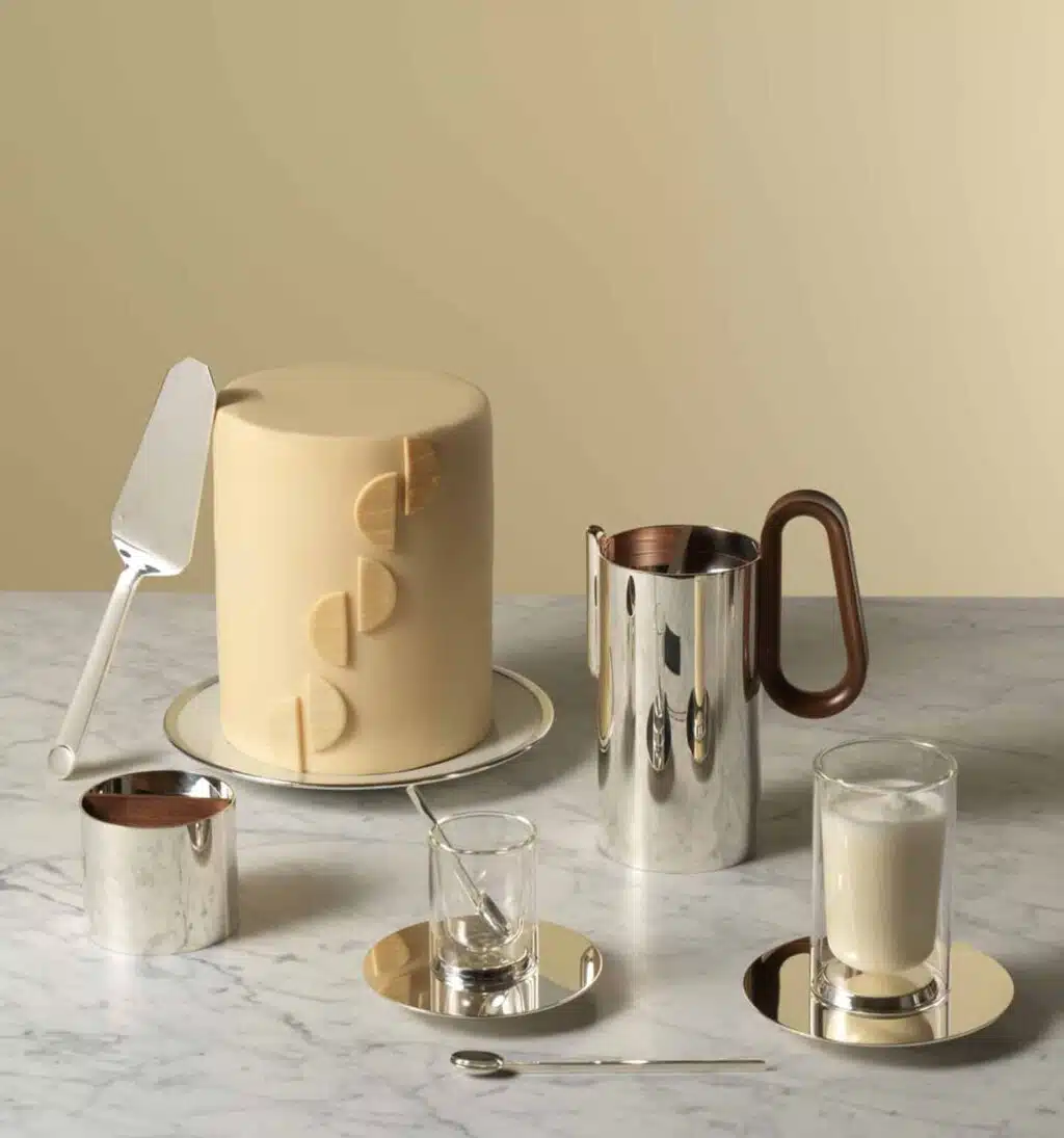 Tea pot, creamer, expresso and infusion cups and saucers photographed from side on a marble table with a big pastry and a cake shovel in front of a pale yellow wall