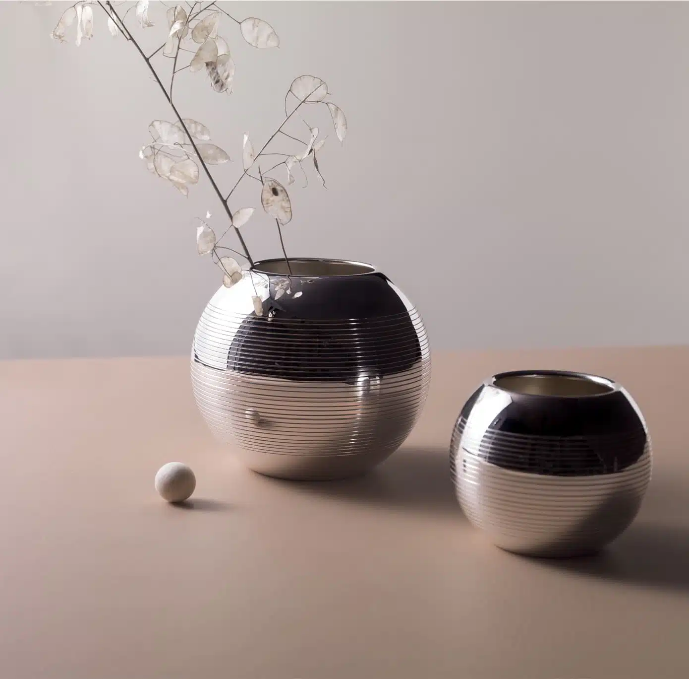 Vases in silver plated from Pétanque collection from Puiforcat photographed on a light pink table in front of a grey wall with a flower in the left vase, and a boule game starter in front